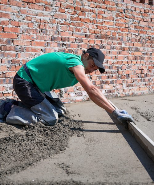 Side view of man in cap placing screed rail on the floor covered with sand-cement mix at construction site. Male worker leveling surface with straight edge while screeding floor in new building.